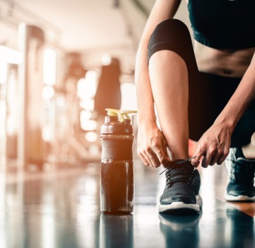 female in black leggings and black tennis shoes crouching on the ground to tie her shoe inside a gym with a black waterbottle next to her