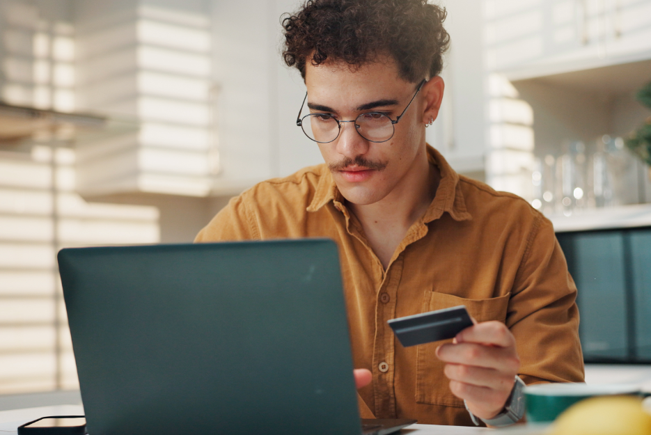 Man with dark curly hair sitting at a laptop with a card in his hand looking to make a payment
